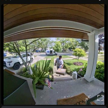 A fisheye security camera view from a covered front porch, showing a person standing near a mailbox, with parked cars and a suburban street visible in the background.