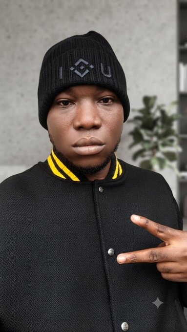 A portrait of a man wearing a black beanie with 'I G U' lettering and a black jacket with a yellow-striped collar, making a peace sign.