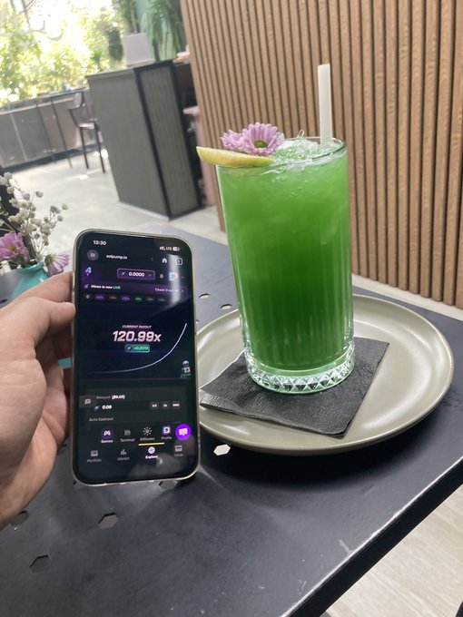 A hand holds a smartphone showing a trading app with a 120.99x leverage position next to a vibrant green cocktail garnished with flowers at an outdoor cafe.