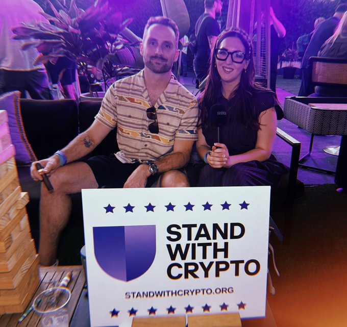A man and a woman sit together holding a 'STAND WITH CRYPTO' sign with the website STANDWITHCRYPTO.ORG, at an event with purple lighting and an RØDE microphone.