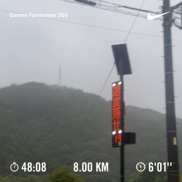 A misty mountain view captured on a Garmin Forerunner 265 during an 8.00 km morning run (48:08, 6'01" pace), with a red Japanese road sign reading "速度落せ" (Slow Down) visible alongside utility poles in the rainy, foggy conditions. The atmospheric scene reflects the runner's post about being defeated by wind and rain during their morning workout.