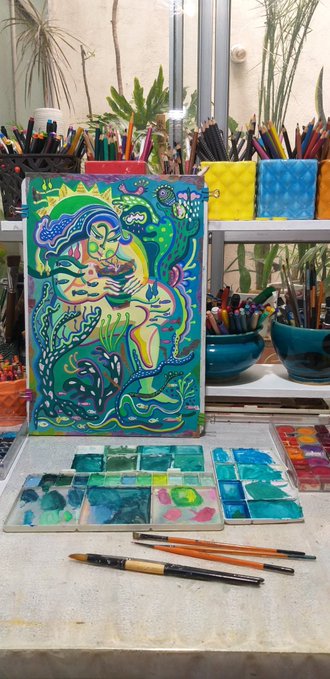 A vibrant blue and green painting of a seated figure surrounded by sea creatures, displayed on an easel in an artist's studio with brushes and watercolors.