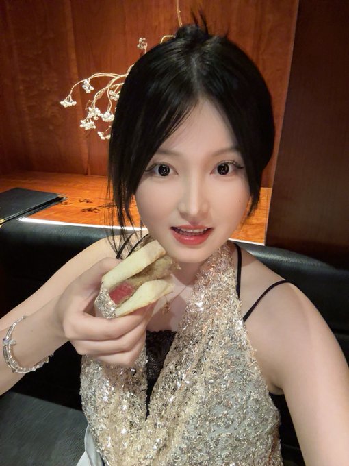 A young woman wearing a sparkly gold sequined top holds a small sandwich, taking a selfie in what appears to be a restaurant.