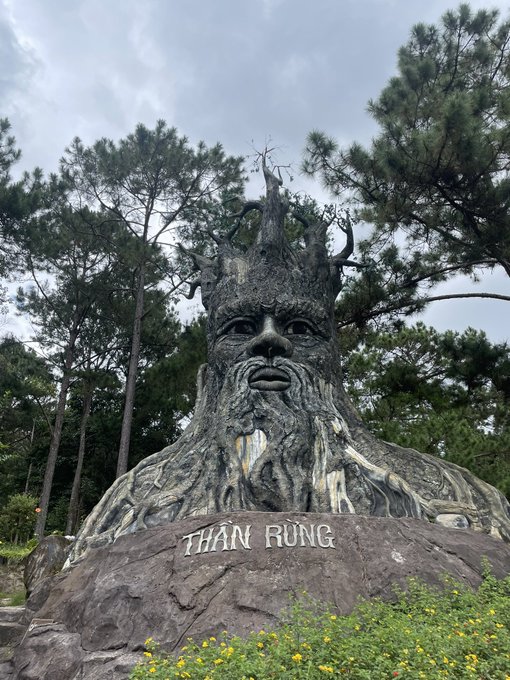 A large stone sculpture of a bearded forest king with tree branches forming his crown, inscribed with 'THẦN RỪNG' (Forest God) on the base, surrounded by pine trees and yellow flowers.