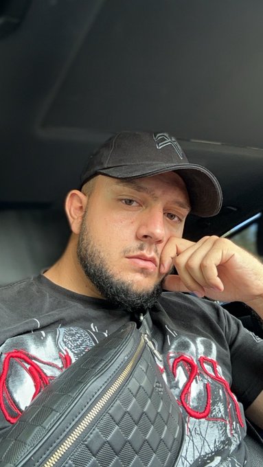 A selfie of a bearded man wearing a black NY Yankees cap and a black graphic t-shirt, sitting inside a car with a crossbody bag.