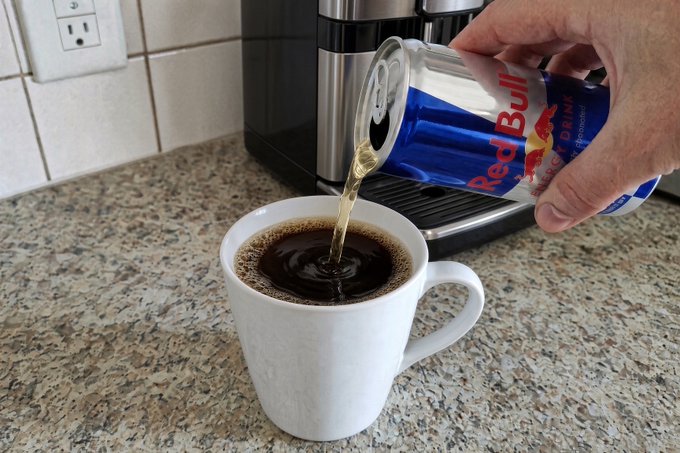 A hand pours a can of Red Bull into a white coffee mug of black coffee, combining the two energy boosters to power an extra "25th hour" of work.