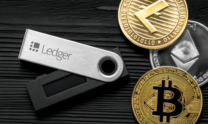 Photo of a Ledger hardware wallet alongside physical representations of Bitcoin, Litecoin, and Ethereum cryptocurrency coins on a black wooden surface.