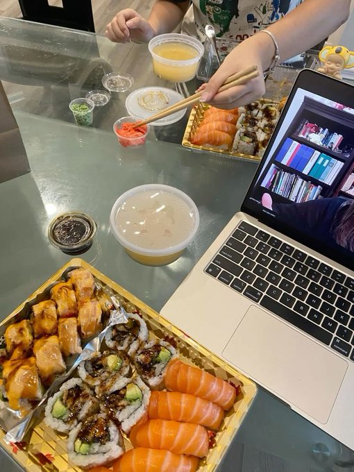 A first-person view of a meal featuring trays of assorted sushi, including salmon nigiri and rolls, alongside a bowl of soup and a laptop on a table.