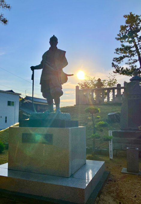 The image features a statue of a man standing next to a building, with the sun setting behind him. The statue appears to be holding a sword, which adds an element of historical significance or artistic expression to the scene. In the background, there are two people present who might be admiring the statue or attending to it.