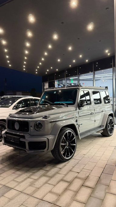 A sleek silver Mercedes-Benz G-Wagon is displayed under showroom lighting, with other luxury vehicles visible in the background. The caption humorously references selling cars like this to invest in Bitcoin ($BTC), implying the post author gained more profit from crypto than holding expensive vehicles.