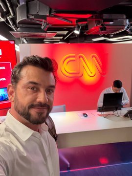 A man with a beard in a white shirt takes a selfie in a studio with red lighting, a glowing "CM" neon sign, and another person working at a computer in the background.
