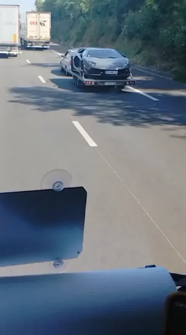 A view from a moving vehicle shows a flatbed tow truck carrying a black sports car down a highway, with trucks visible in the distance.