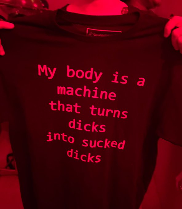 A person holds up a dark t-shirt with red text reading "My body is a machine that turns dicks into sucked dicks" under red lighting.