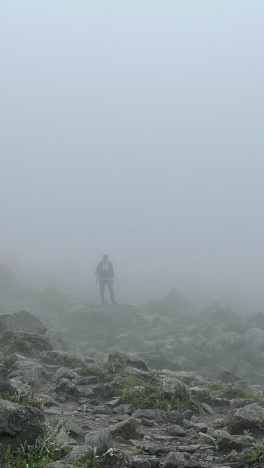 A lone figure with a backpack stands on rocky, mist-shrouded terrain enveloped in dense fog, with sparse vegetation dotting the foreground.