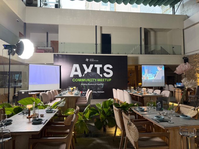 The image shows an indoor event space set up for the "AXIS ROBOTICS COMMUNITY MEETUP" in Ho Chi Minh City, Vietnam, with elegant dining tables, tropical plants, and a large screen displaying the event branding. The venue arrangement reflects a sophisticated gathering where attendees from the Axis Robotics community could meet, network, and exchange ideas face-to-face as referenced in the post.