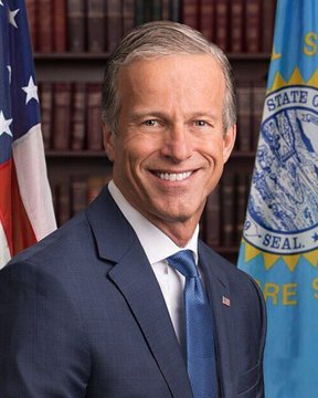 The image shows a smiling man in a blue suit and tie, standing in front of an American flag and a U.S. Senate flag, with bookshelves in the background—appearing to be an official portrait of Senator John Thune of South Dakota, who is referenced in the post about pushing the CLARITY Act crypto legislation review to September.