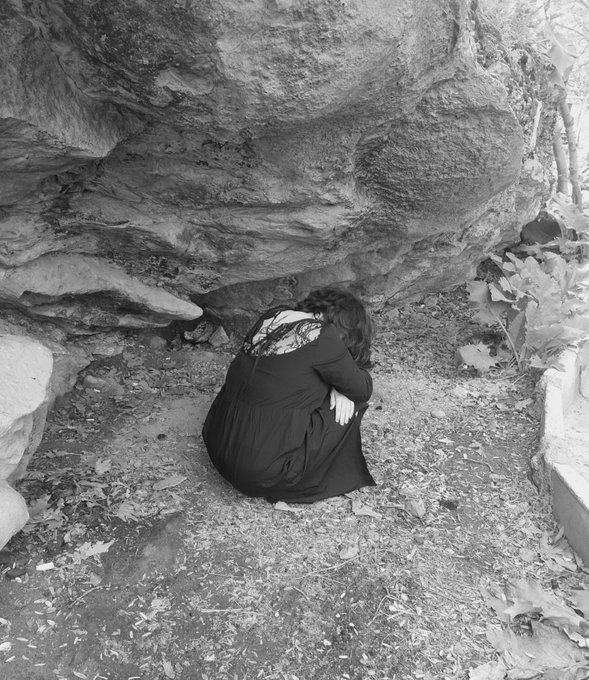 A black and white photograph of a woman in a dark dress sitting on the ground with her knees pulled up, leaning against a large rock face in a natural setting.