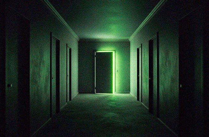 A dark, eerie hallway illuminated by a bright green light spilling through an open door at the end, with several closed doors lining the walls.