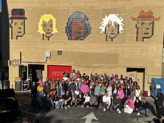 A large group of people poses in front of a tan building adorned with pixel-art murals of five faces wearing various hats, with a partially visible "NODE" sign on the red door behind them, celebrating the Node Foundation's accomplishments in 2026.