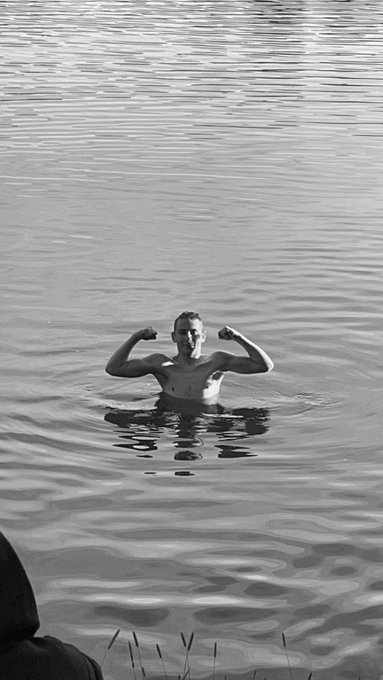 A black-and-white photo shows a man standing waist-deep in water, flexing both biceps toward the camera.