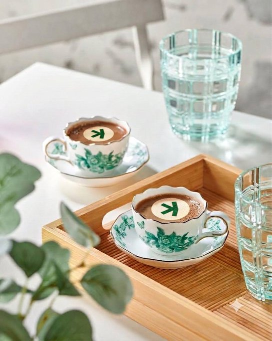 Two green and white floral-patterned teacups filled with frothy coffee drinks (each topped with a green chevron logo) on a wooden tray, accompanied by two textured green glass tumblers and eucalyptus leaves.