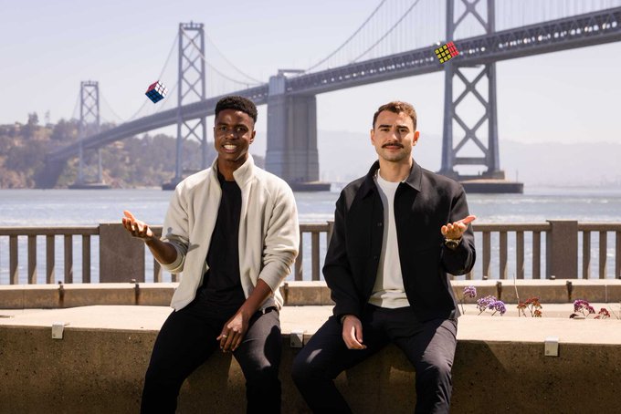 Two men sit casually on a concrete barrier near a waterfront with a large suspension bridge in the background, each tossing small Rubik's cubes in the air as they smile and gesture toward the camera, reflecting the post's theme of playful problem-solving for AI agents at os.virtuals.io.