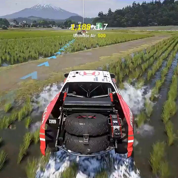 The image shows a red and white truck driving through a field, with water splashing around its tires as it moves across the landscape. The truck appears to be racing or participating in an off-road event, given that it has large tires on its back. In the background, there is a mountain range visible behind the truck, adding to the sense of adventure and excitement associated with this activity.