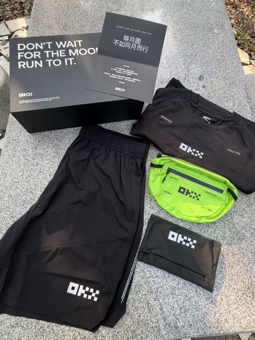 An OKX running festival box reading 'DON'T WAIT FOR THE MOON. RUN TO IT.' with black shorts, a t-shirt, a neon green belt bag, and a wristband laid on stone.