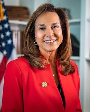 The image features a woman wearing a red jacket, smiling at the camera. The caption reads "LATEST: According to CNBC, Rep. Lisa McClain's family could reportedly see a six-figure gain from an early xAI investment that became SpaceX shares ahead of the company’s IPO." This statement suggests that there is a connection between the woman in the red jacket and a recent event involving Rep. Lisa McClain and her family's financial gains due to an early AI investment by SpaceX.