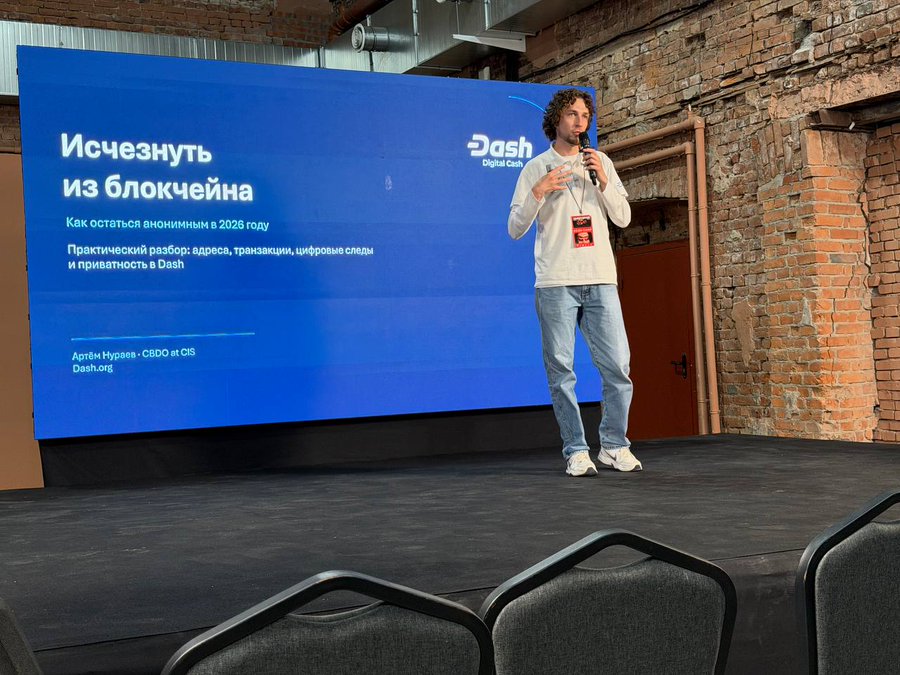 A man with curly hair speaking on a stage at a conference, with a large screen behind him displaying a presentation in Russian titled 'Исчезнуть из блокчейна' (Disappear from the blockchain) about remaining anonymous in 2026, featuring the Dash Digital Cash logo and the speaker's name, Артём Кураев.