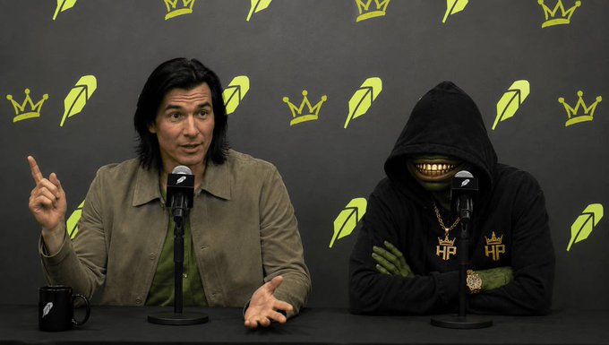 A man in a green shirt and tan jacket speaking into a microphone next to a green-skinned, sharp-toothed creature in a black hoodie, with a dark background patterned with yellow crowns and feathers.