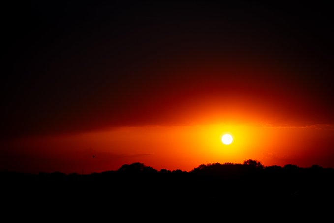 A scenic photograph of a bright yellow sun setting against a deep red and black sky, silhouetting a distant tree line.