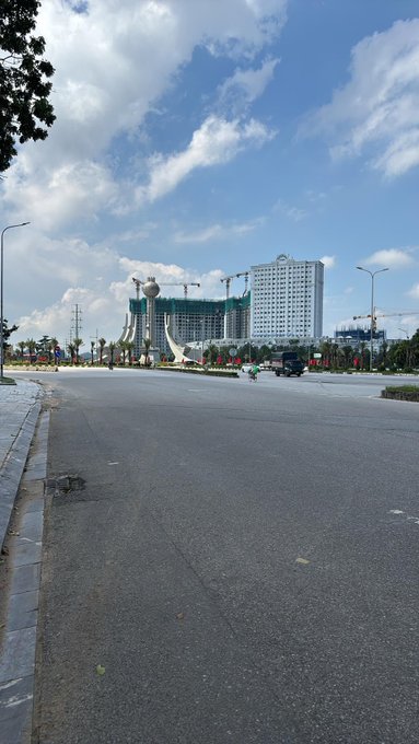 A wide empty street view with a tall white hotel/building and construction cranes in the background, under a partly cloudy blue sky.