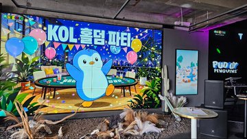 A vibrant event space features a large illuminated display reading "KOL 홀덤 파티" alongside a cheerful penguin mascot and colorful balloon graphics, with a "Pudgy Penguins" branded booth visible on the right. The festive setup, complete with tropical plants and decorative tables, reflects the lively atmosphere of the Winners KOL Hold'em Party Meetup hosted at the Weple NFT gallery in collaboration with Pudgy Penguins.