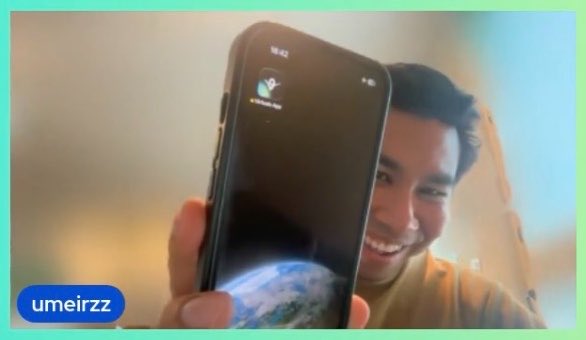 A man smiles behind a smartphone displaying the "Virtuals Bot" app, with a username "umeirzz" in the corner, capturing the moment of engaging with the @virtuals_io platform.