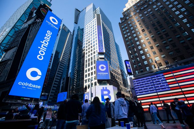 A crowd gathers in an urban plaza beneath towering digital billboards prominently displaying the Coinbase logo and the iconic Bitcoin "₿" symbol against the backdrop of a skyscraper. The Coinbase branding reinforces the company's presence in the crypto and financial markets, aligning with its announcement of expanding IPO access to U.S. retail customers.
