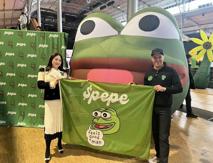 Two people holding a green $pepe banner in front of a giant inflatable Pepe frog and a yellow flower at what appears to be a convention.