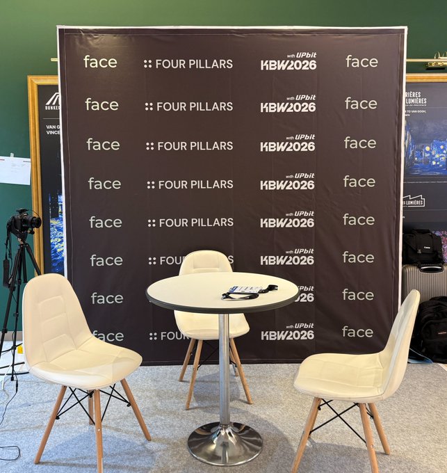 An event booth setup with a black backdrop displaying 'face', 'FOUR PILLARS', and 'KBW2026' logos, featuring a small round table and three white chairs.