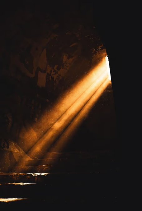 Sunlight streams through a small opening in a dark cavern, illuminating golden dust particles as the light rays slant downward into the rocky interior.