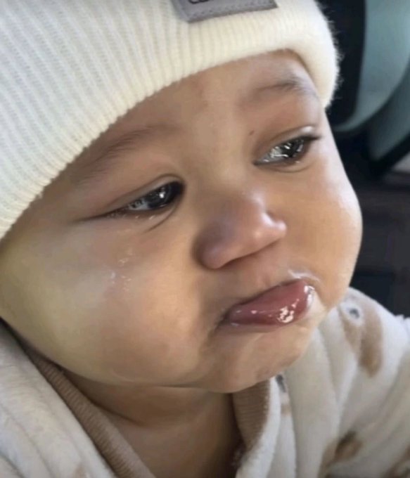 A close-up of a baby wearing a white beanie, looking at the camera with a tear on their cheek.