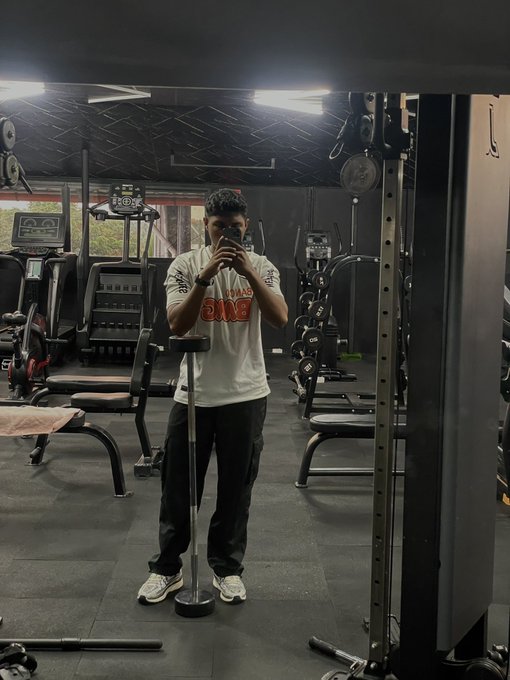 A man in a white graphic t-shirt and black pants takes a mirror selfie in a gym while holding a smartphone on a tripod.