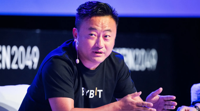 A man in a black t-shirt speaks at a conference, wearing a microphone headset, with a backdrop showing the text 'TOKEN2049' and a shirt logo reading 'BYBIT'.
