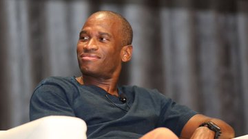 The image features an older man wearing a blue shirt sitting down with his legs crossed. He has a serious expression as he looks off into the distance. The background of the photo shows a curtain, which adds depth to the scene. There are also some words visible in the image that seem to be related to Arthur Hayes and his recent transactions on cryptocurrency.