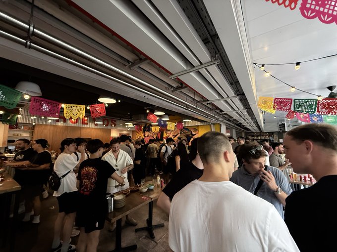 A crowded restaurant interior decorated with colorful papel picado banners, featuring many patrons standing around tables.