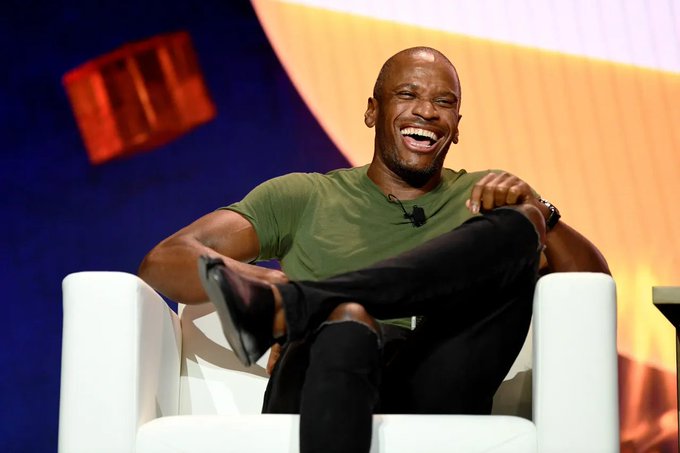 The image features a man sitting in a white chair with his legs crossed, smiling as he looks directly at the camera. The background of the photo shows an orange wall with a large red box behind him. There are also some text elements present in the image that read "Arthur Hayes just bought back 33,978 $HYPE after turning super bearish on it just a few days ago."