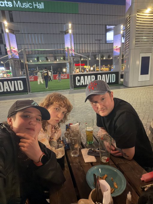 The image shows a group of three people sitting at an outdoor table, enjoying their time together. They are posing for a picture with smiles on their faces. The table has various items on it, including cups, bowls, forks, and knives. There is also a cell phone placed on the table. In the background, there is a building that appears to be a music hall or stadium, suggesting they might have attended an event at this venue.