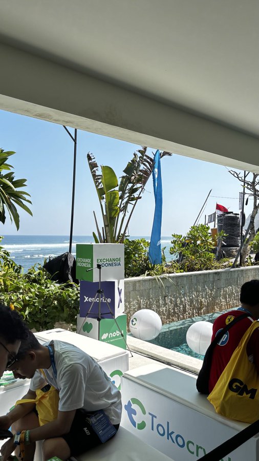 A scene at what appears to be a crypto/tech event with a sign reading 'EXCHANGE INDONESIA' and 'Xendit' on a podium with a tripod, set against an ocean view with people sitting nearby and TokoCrypto branding visible.
