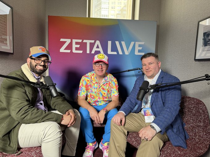 Three men sit in armchairs with microphones for a podcast recording in front of a colorful “ZETA LIVE” backdrop; the center guest wears a bright patterned shirt and colorful cap, while the host on the right wears a blue blazer and a “ZETA LIVE” badge. The image matches the post’s mention of a conversation with Dan from Basis Point Podcast at Zeta Live.
