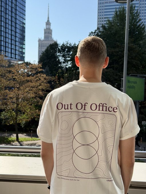 A man stands facing away from the camera, wearing a white t-shirt with the text 'Out Of Office' and a topographic pattern design featuring interlocking circles, with the Palace of Culture and Science visible in the background.