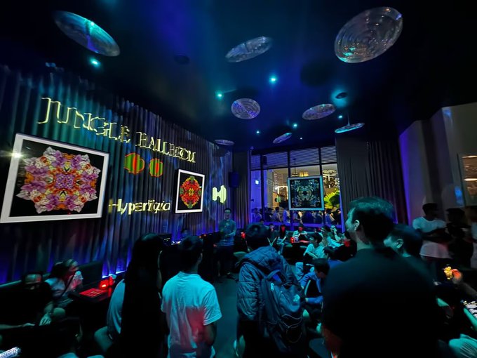 A crowded, dimly lit event space features a 'JUNGLE BALLROOM' sign on the wall alongside colorful artwork and 'Hyperfy' branding, with attendees mingling under circular ceiling lights.